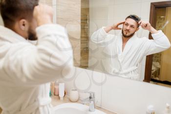 Man in bathrobe combs his hair in bathroom, routine morning hygiene. Male person at the sink performs skin and body treatment procedures