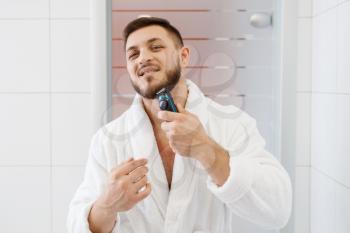 Man in bathrobe shaves his beard with an electric razor in bathroom, routine morning hygiene.