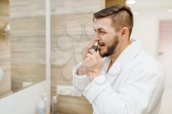 Man in bathrobe removes nose hair in bathroom, routine morning hygiene. Male person at the sink performs skin and body treatment procedures