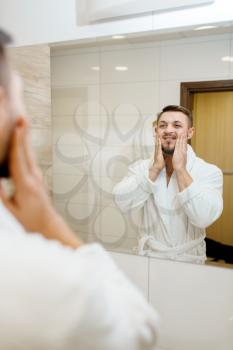 Man in bathrobe rubs aftershave over his face at the mirror in bathroom, routine morning hygiene. Male person at the sink performs skin and body treatment procedures