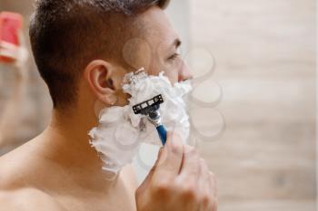 Man shaves his beard with razor in bathroom, routine morning hygiene. Male person at the sink performs skin and body treatment procedures