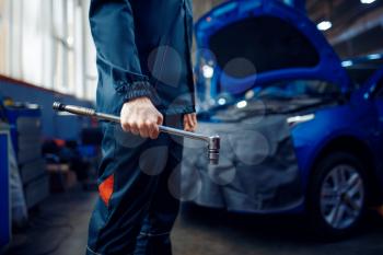 Worker in uniform holds wrench, vehicle with opened hood on background, car service station. Automobile checking and inspection, professional diagnostics and repair
