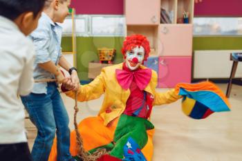 Funny clown with joyful children playing tug of war together. Birthday party celebrating in playroom, baby holiday in playground. Childhood happiness, childish leisure