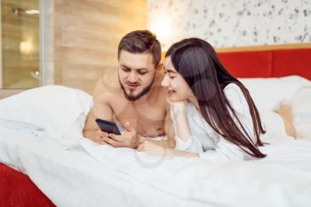 Love couple in pajamas using phone in bed at home, good morning. Harmonious relationship in young family. Man and woman resting together in their house, carefree weekend
