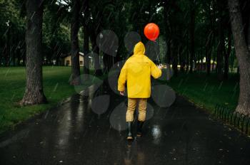Sad man with balloon walking in summer park in rainy day. Male person in rain cape and rubber boots, wet weather in alley, loneliness
