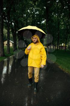 Man with umbrella walking in summer park in rainy day. Male person in rain cape and rubber boots, wet weather in alley
