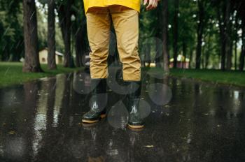 Male person in rain cape and rubber boots, wet weather in alley. Man poses in summer park in rainy day. Water protection, drops