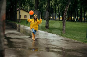 Happy woman runs with balloon in summer park in rainy day. Female person in rain cape and rubber boots, wet weather in alley, loneliness