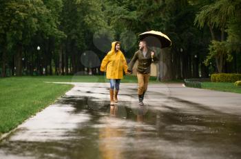 Happy love couple runs in park, summer rainy day. Man and woman under umbrella in rain, romantic date on walking path, wet weather in alley