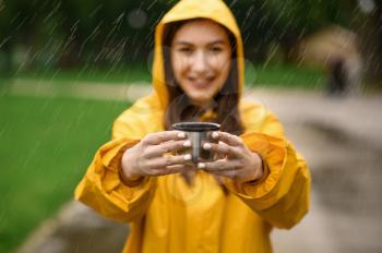 Woman in raincoat holds cup of hot tea, summer park in rainy day, view through the drops. Alone female person in rain cape on walking path, wet weather in alley