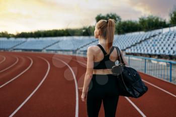Female runner in sportswear holds sport bag, back view, training on stadium. Woman doing stretching exercise before running on outdoor arena