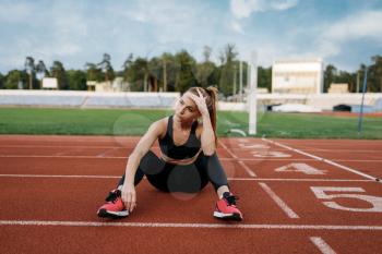 Tired female jogger sitting on the ground, training on stadium. Woman doing stretching exercise before running on outdoor arena