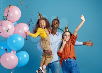 Happy family celebrate birthday, blue background. Little girl and her parents in caps blowing party whistles, balloons and confetti decoration