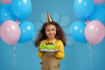 Funny little girl in cap holds birthday cake, blue background. Pretty child got a surprise, event celebration, balloons decoration