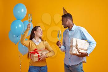 Funny couple in caps holding glasses of beverage and gift boxes, yellow background. Pretty female person got a surprise, event or birthday celebration, balloons decoration