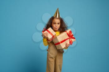 Funny little girl in cap holds birthday gift box with red ribbons, blue background. Pretty child got a surprise, event celebration