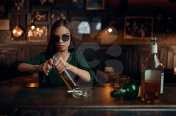Woman in sunglasses opens bottle of beer at the counter in bar. One female person in pub, human emotions, leisure activities, depression