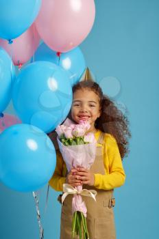 Funny little girl in cap holds a bunch of colorful balloons and flower bouquet, blue background. Pretty child got a surprise, event or birthday party celebration