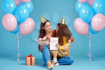 Funny little girl congratulates her mother, blue background. Pretty child hugs her mom, event or birthday party celebration, balloons and gift box decoration
