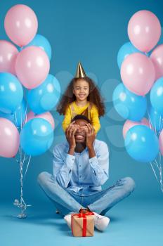 Funny female kid congratulates her father, blue background. Pretty child hugs her dad, event or birthday party celebration, balloons and gift box decoration