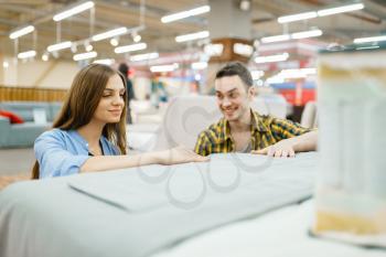Young happy couple buying blanket for bed in furniture store. Man and woman looking samples for bedroom in shop, husband and wife buys goods for modern home interior