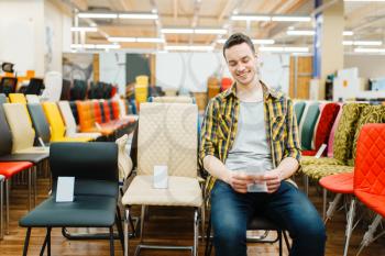 Young man sitting on chair in furniture store showroom. Male person looking seats in shop, husband buys goods for modern home interior