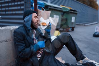 Bearded dirty beggar with food sitting at the trashcan on city street. Poverty is a social problem, homelessness and loneliness, alcoholism and drunk addiction