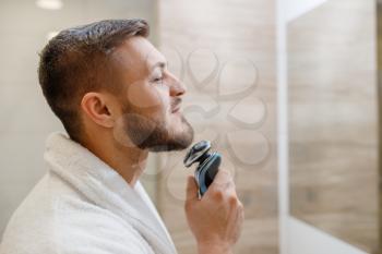 Man at the mirror shaves his beard with an electric razor in bathroom, routine morning hygiene. Male person at the sink performs skin and body treatment procedures