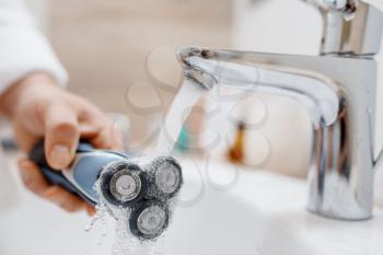 Man cleans electric razor after shaving in bathroom, routine morning hygiene. Male person at the sink performs skin and body treatment procedures