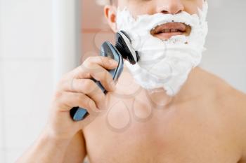 Man shaves his foamed beard with an electric razor in bathroom, routine morning hygiene. Male person at the sink performs skin and body treatment procedures
