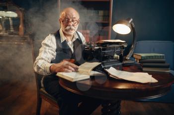 Elderly writer works on vintage typewriter in his home office. Old man in glasses writes literature novel