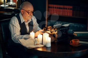 Adult writer works on vintage typewriter with candle light in home office. Old man in glasses writes literature novel in room with smoke, inspiration