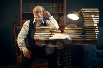 Elderly writer sitting at the table with stack of books in home office. Old man in glasses writes literature novel in room with smoke, inspiration