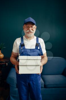 Elderly cargo man in uniform poses in home office. Adult delivery worker, deliver in cap holds boxes indoors, delivering service or business