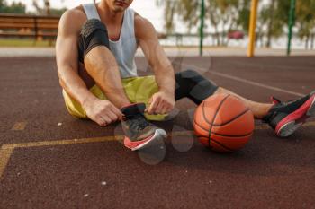 Basketball player sitting on the ground and tying laces on outdoor court. Male athlete in sportswear resting after streetball training