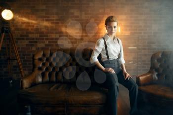 Sexy business woman in strict clothes poses on leather couch in studio, retro fashion, gangster style, mafia. Vintage lady in office with brick walls