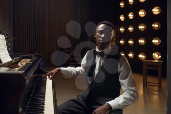 Ebony grand piano player on the stage with spotlights on background. Negro performer poses at musical instrument before concert