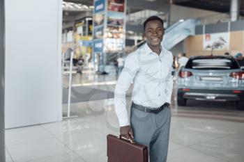 Smiling businessman holds briefcase in car showroom. Successful business person on motor show, black man in formal wear