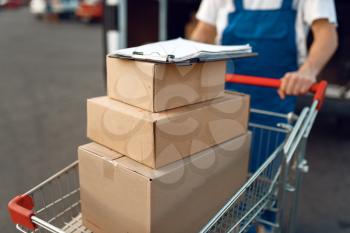 Deliveryman in uniform holds cart with stack of boxes at the car with parcels, delivery service. Man standing at cardboard packages in vehicle, male deliver, courier or shipping job