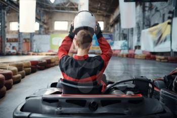 Kart racer puts on helmet, back view, karting indoor. Speed race on close go-cart track with tire barrier. Fast vehicle competition, high adrenaline leisure