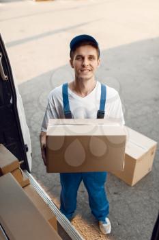 Deliveryman in uniform unloads the car with parcels, delivery service. Man standing at cardboard packages in vehicle, male deliver, courier or shipping job
