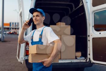 Deliveryman in uniform holds parcel and mobile phone at the car, delivery service, delivering. Man standing at cardboard packages in vehicle, male deliver, courier or shipping job