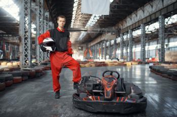 Racer with helmet poses near go kart car, karting auto sport indoor. Speed race on close go-kart track with tire barrier. Fast vehicle competition, high adrenaline leisure