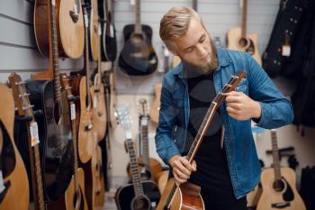 Bearded young man choosing acoustic guitar in music store. Assortment in musical instruments shop, male musician