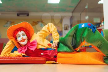 Funny clown in costume lying on the floor in children's area. Birthday party celebrating in playroom, baby holiday in playground. Childhood happiness, childish leisure with animator