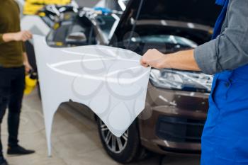 Male workers holds front fender template of car protection film. Installation of coating that protects the paint of automobile from scratches. New vehicle in garage, tuning procedure