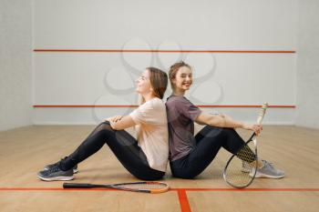 Two female players with squash rackets sits back to back on court floor. Girls on training, active sport hobby, fitness workout for healthy lifestyle