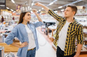 Young couple jokes with ladle and pan in houseware store. Man and woman buying home goods in market, family in kitchenware supply shop
