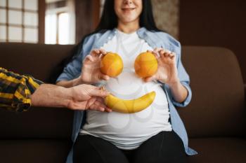 Couple jokes with fruits, husband and his pregnant wife with belly relaxing at home. Pregnancy, prenatal period. Expectant mom and dad are resting, health care