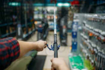 Male angler with rod in fishing shop, first-person view. Equipment and tools for fish catching and hunting, accessory choice on showcase in store, spinnings and telescopes assortment
